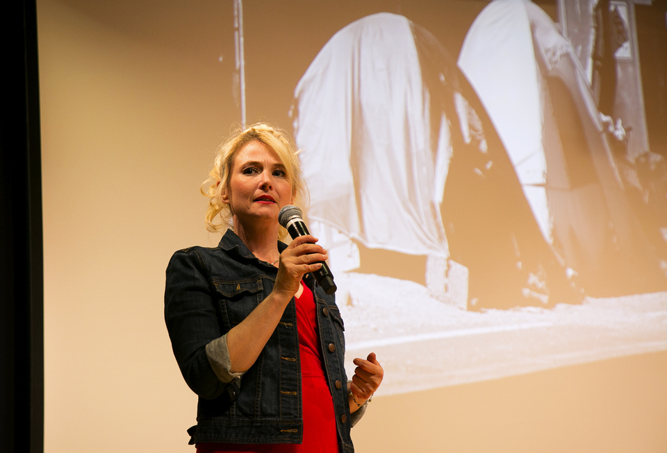 Woman Wearing Jean Jacket And Red Shirt Standing And Talking In Microphone With Images Of Seattle Homeless Tents In Background