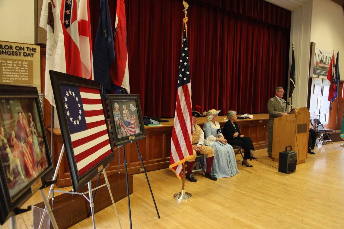 Man In Brown Suit Standing In Front Of Podium Talking In A Hall With People Dressed As Pilgrims