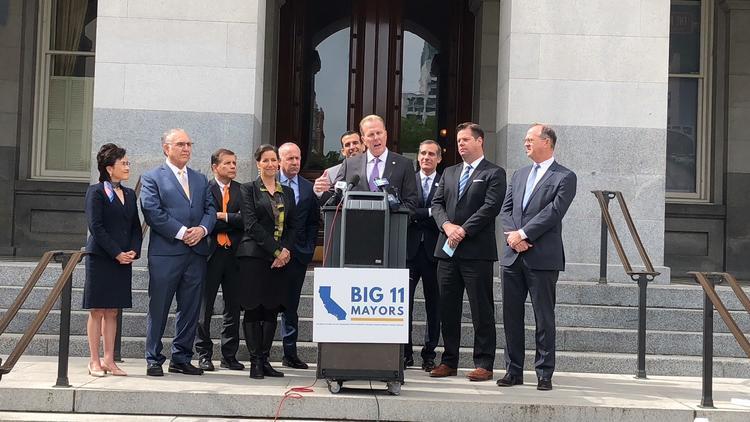 Man Wearing Suit And Tie Standing In Front Of Podium With Microphones With Big 11 Mayor Sign In San Diego With Group Of People In Suits In Background