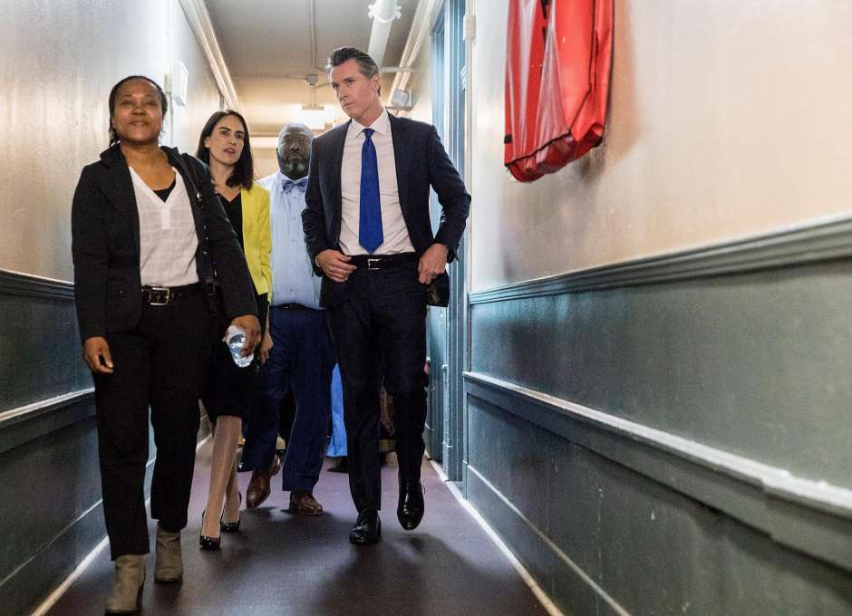 Governor Gavin Newsom Walking Down Green And White Hallway With Three Other People Discussing California Homelessness